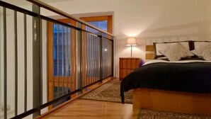 A bedroom with a bed, a nightstand, a lamp, and a view of the outside through a window. - Central Mezzanine Apartment in Historic School (Edinburgh)
