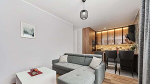 A modern living room with a grey sectional sofa, a small white side table, and a kitchen area with wooden cabinets and bar stools. - Stylish Apartment with Balcony | Parking | Close to the Oder River (Wroclaw)