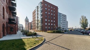 A modern urban street with a mix of residential and commercial buildings, a parking lot, and a grassy area with a bench. - Stylish Apartment with Balcony | Parking | Close to the Oder River (Wroclaw)