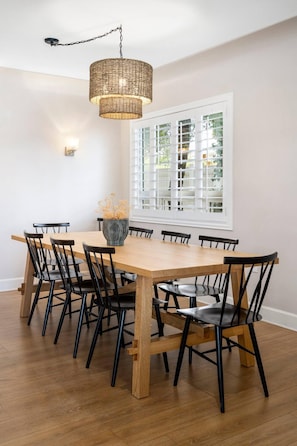 Un coin repas avec une table en bois et des chaises noires, un luminaire suspendu en fibres tressées et une fenêtre avec des stores.