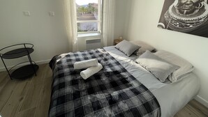 Un dormitorio con una cama, un ventanal, un cuadro colgado en la pared y una mesita.