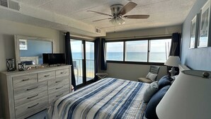 A bedroom with a bed, a dresser, a television, a ceiling fan, and a view of the ocean through large windows. - Spacious, comfortable 2 bedroom oceanfront condo with free WiFi, an outdoor pool, and an enclosed balcony with a great ocean view located midtown only steps to the beach! (Ocean City)
