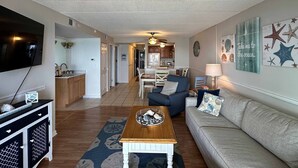 A living room with a TV, sofa, coffee table, and kitchen in the background. - Spacious, comfortable 2 bedroom oceanfront condo with free WiFi, an outdoor pool, and an enclosed balcony with a great ocean view located midtown only steps to the beach! (Ocean City)