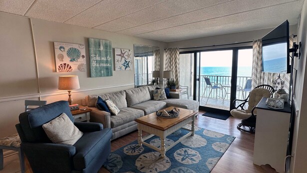 A living room with a sofa, armchair, coffee table, and a view of the sea through large glass doors. - Spacious, comfortable 2 bedroom oceanfront condo with free WiFi, an outdoor pool, and an enclosed balcony with a great ocean view located midtown only steps to the beach! (Ocean City)