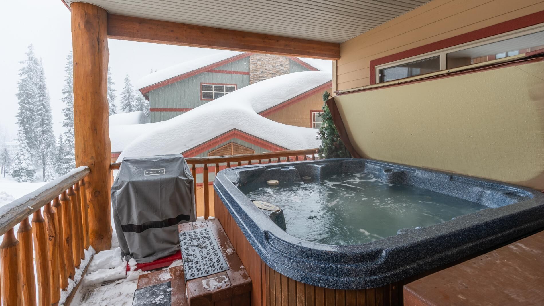 A hot tub on a wooden deck with a covered grill and a snow-covered house in the background.