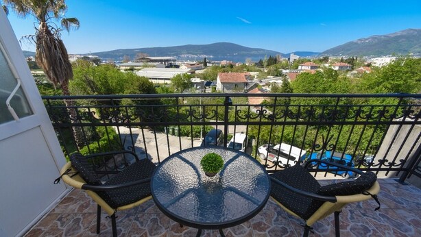 Un balcon avec deux chaises et une table, d’où l’on peut admirer une ville avec ses bâtiments et, au loin, des montagnes.