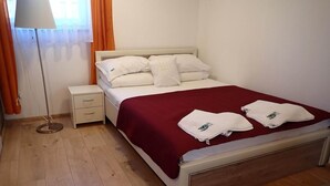 A neatly made bed with white linens and a red blanket, a bedside table with a lamp, and a window with orange curtains.