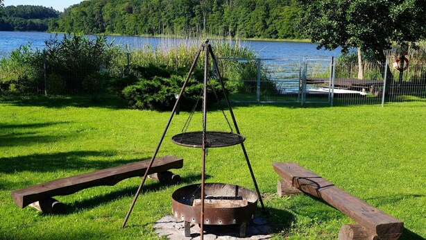 A metal fire pit with a hanging grill, surrounded by a grassy area with picnic benches.