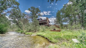 A rustic cabin with a stone foundation and a wooden deck, surrounded by dense forest and a clear stream. - Gorgeous Rippling Waters!  Private Hot Tub- Spectacular Views! (Nathrop)