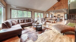 A spacious living room with a large sectional sofa, a round coffee table, and a leather ottoman. The room features wooden flooring, large windows, and a view of the kitchen area. - Gorgeous Rippling Waters!  Private Hot Tub- Spectacular Views! (Nathrop)