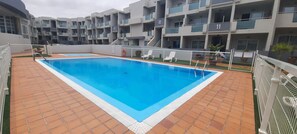 Une piscine dotée d’une terrasse carrelée, entourée d’une clôture blanche et jouxtant un immeuble à plusieurs étages avec des balcons.