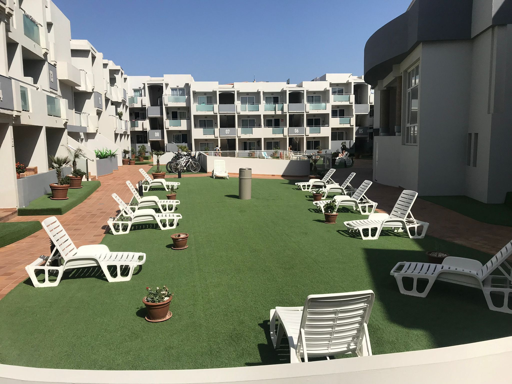Un patio con sillones blancos dispuestos sobre césped artificial, rodeado por un edificio de varios pisos con balcones.