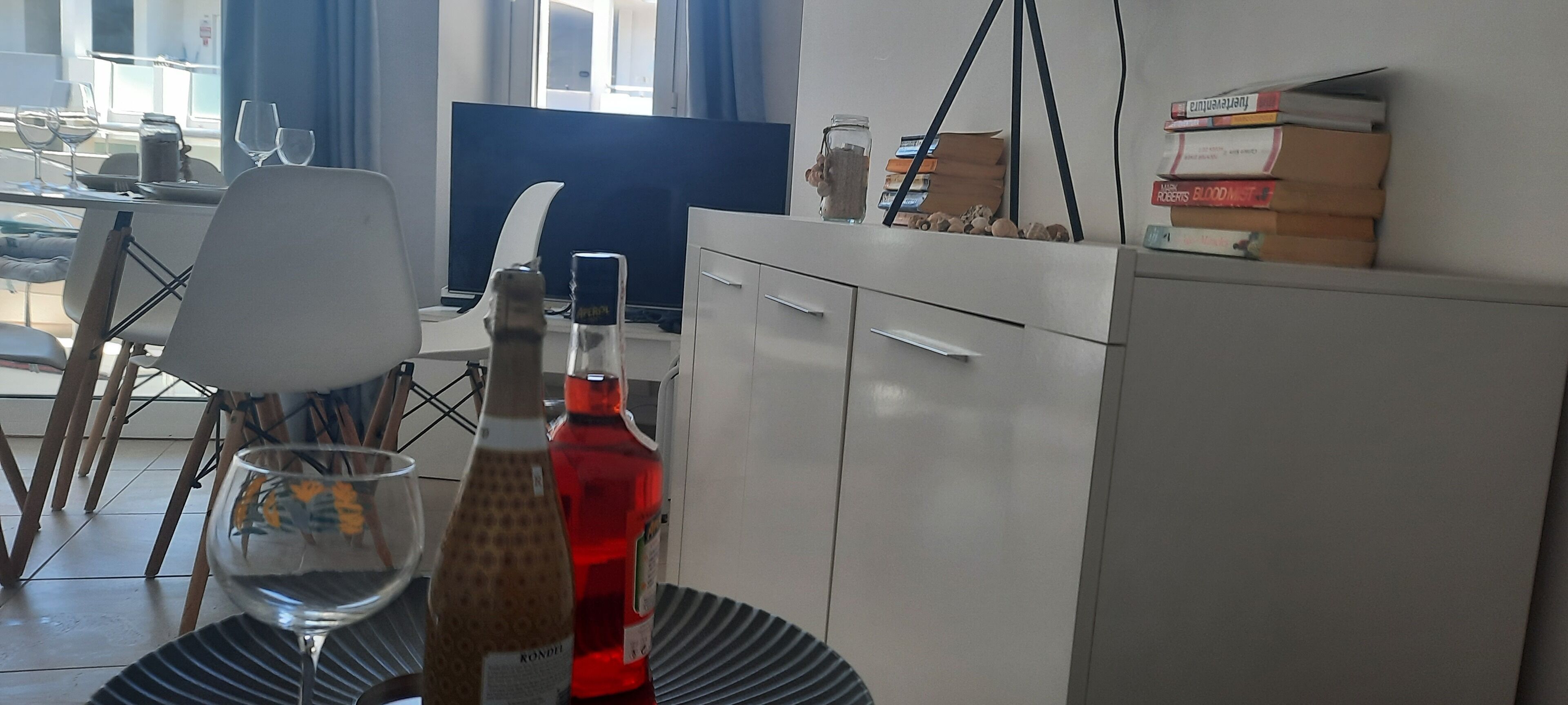 A dining area with a table set for two, a bottle of red wine, and a glass of water. There is a white cabinet with drawers and a blue chair.