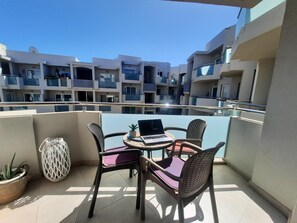 Un balcon avec une table ronde, deux chaises en osier, un ordinateur portable, une plante en pot et un panier tressé.