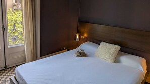 A hotel room with a bed, a nightstand, a lamp, and a view of greenery outside. - Sunrise: Elegant 2BR/1BA in the Heart of Central Barcelona, Dreta de l'Eixample (Barcelona)