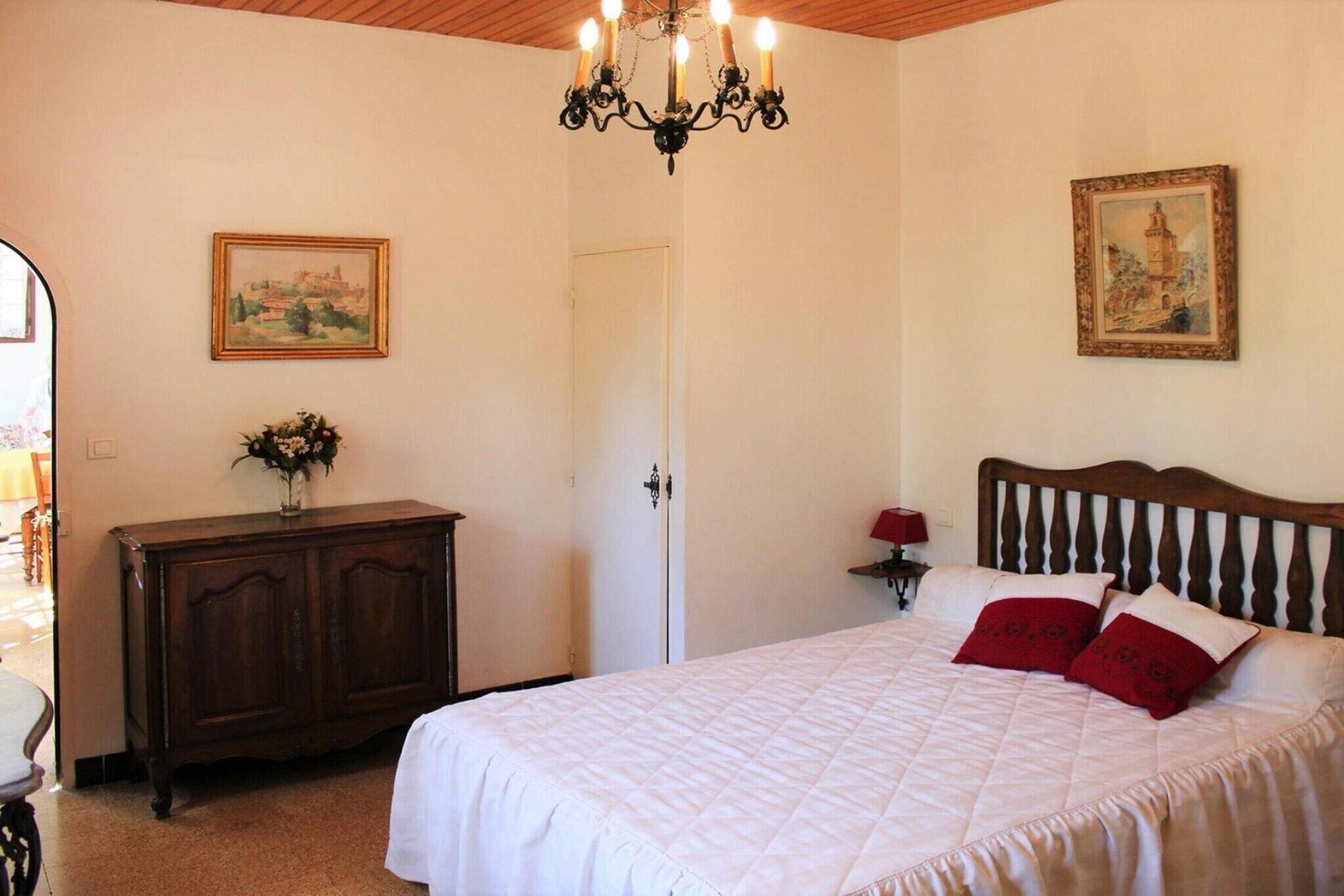 A bedroom with a large bed, a wooden headboard, a chandelier, a dresser, and two framed pictures on the walls.