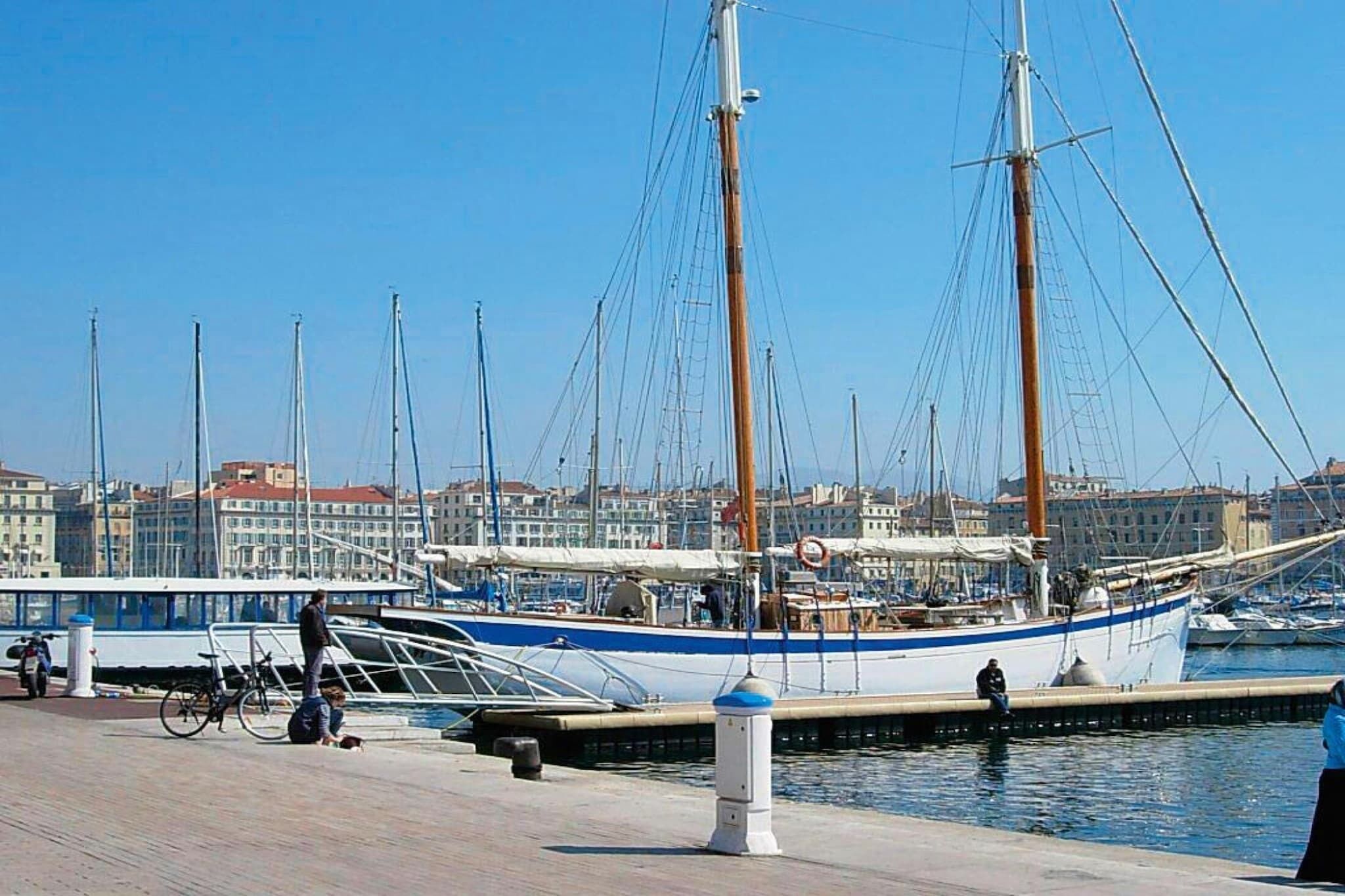 Una marina con varios veleros amarrados, un paseo peatonal y edificios al fondo.