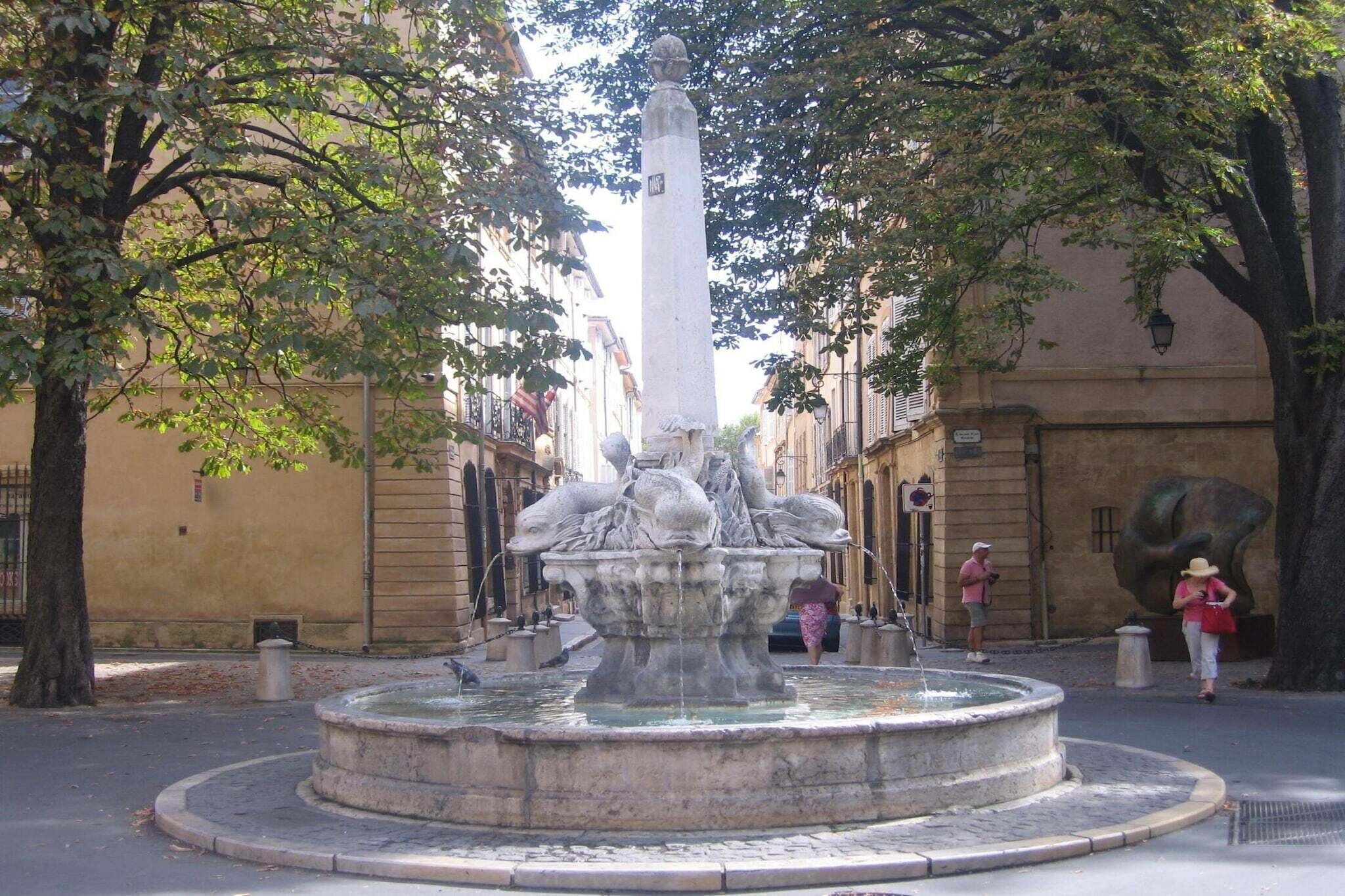 A historic fountain with a central obelisk, surrounded by trees and buildings.