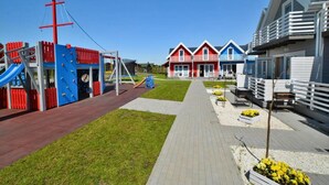 Een speeltuin met klimconstructies en glijbanen, een pad dat leidt naar een rij kleurrijke huizen, en een gazon met bloembedden.