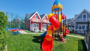 Een speeltuin met een glijbaan en klimconstructies, omringd door huizen met rode daken en witte details.