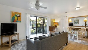En stue med fjernsyn, sofa, spisebord, stole og en balkon med udsigt.