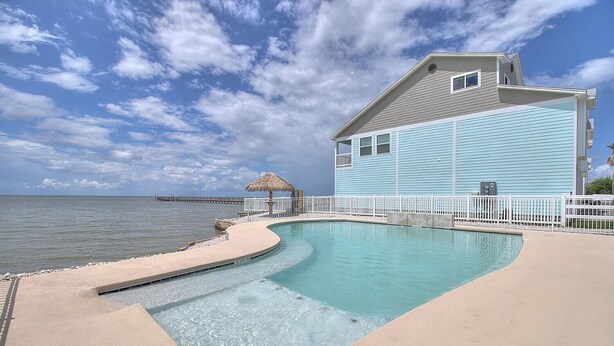 A pool with a curved edge, a thatched-roof structure, and a light blue building with a white fence.
