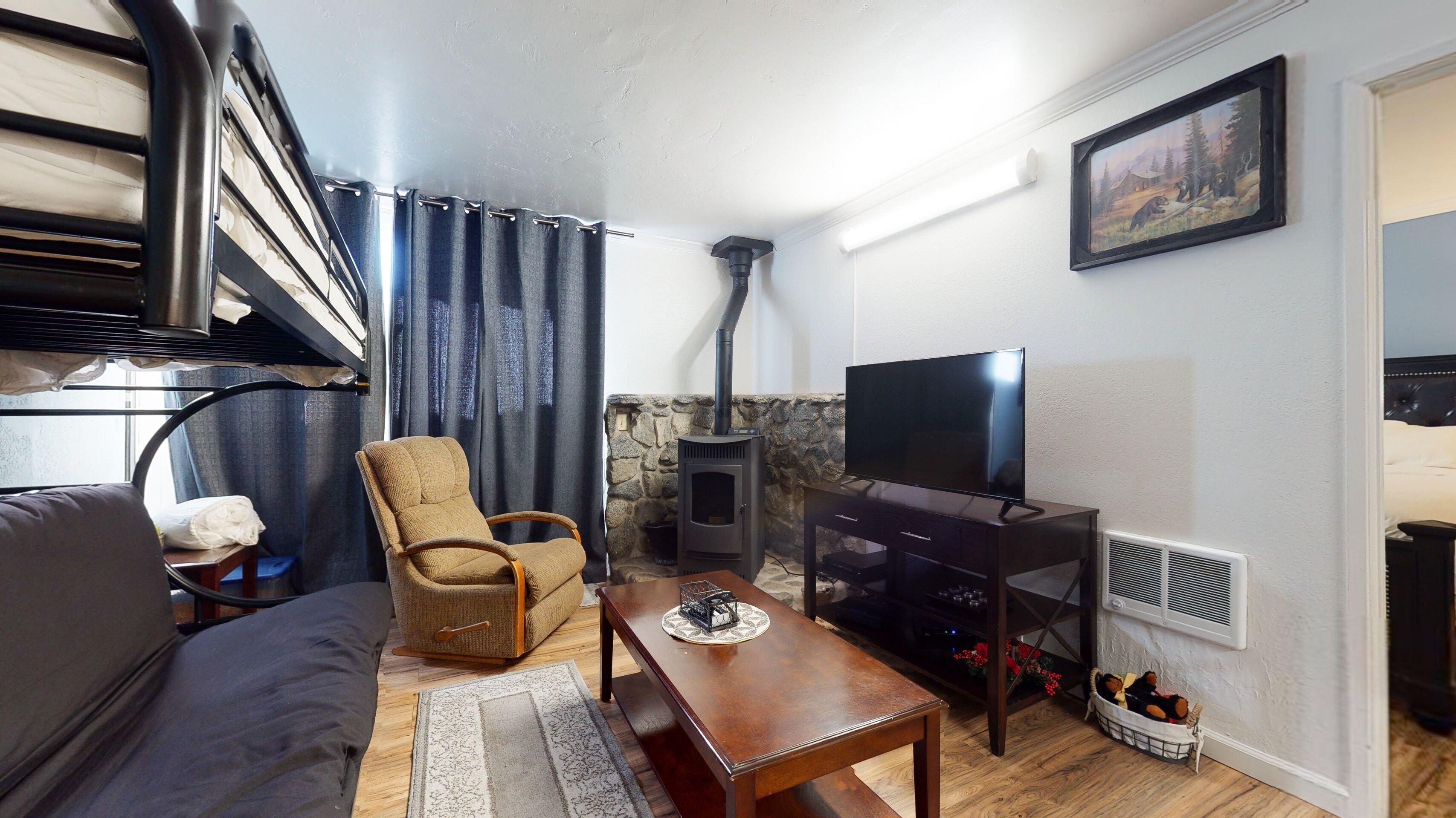 A cozy living room with a sofa, armchair, wooden coffee table, and a TV. A stone fireplace is in the corner, and there's a painting on the wall.