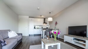 A modern living room with a grey sofa, a coffee table with flowers, a TV, and a kitchen in the background. - Spacious and luxurious apartment - Kaag Resort (22) (Warmond)