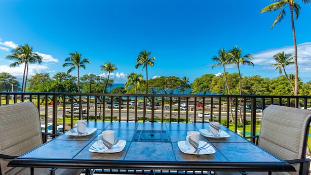 A table set for two with a view of palm trees and a clear sky. - Maui Parkshore #412 Ocean View in the Heart of Kihei, Walk to Kamaole Beach (Kihei)