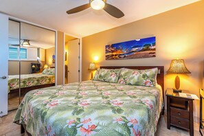 Un dormitorio con una cama grande, un ventilador de techo, una mesita de noche con lámpara, un cuadro en la pared y un espejo en la habitación contigua.