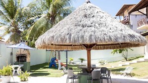 Un espace patio agrémenté d’un parasol en chaume, de sièges d’extérieur, d’un four en briques et d’un toboggan pour les enfants.