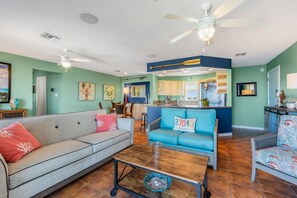 A living room with a sofa, a coffee table, and a blue couch. There is a kitchen in the background with a bar area.