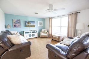 Uma sala de estar com um sofá de couro marrom, um armário branco, cadeiras de vime e um ventilador de teto.