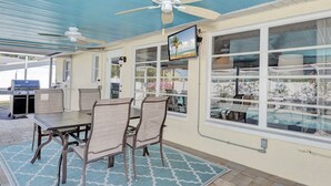 An outdoor patio with a table and chairs, a ceiling fan, and a TV screen displaying a beach scene. - Private heated pool. Shark teeth, close to beaches, downtown, shopping, biking… (Venice)