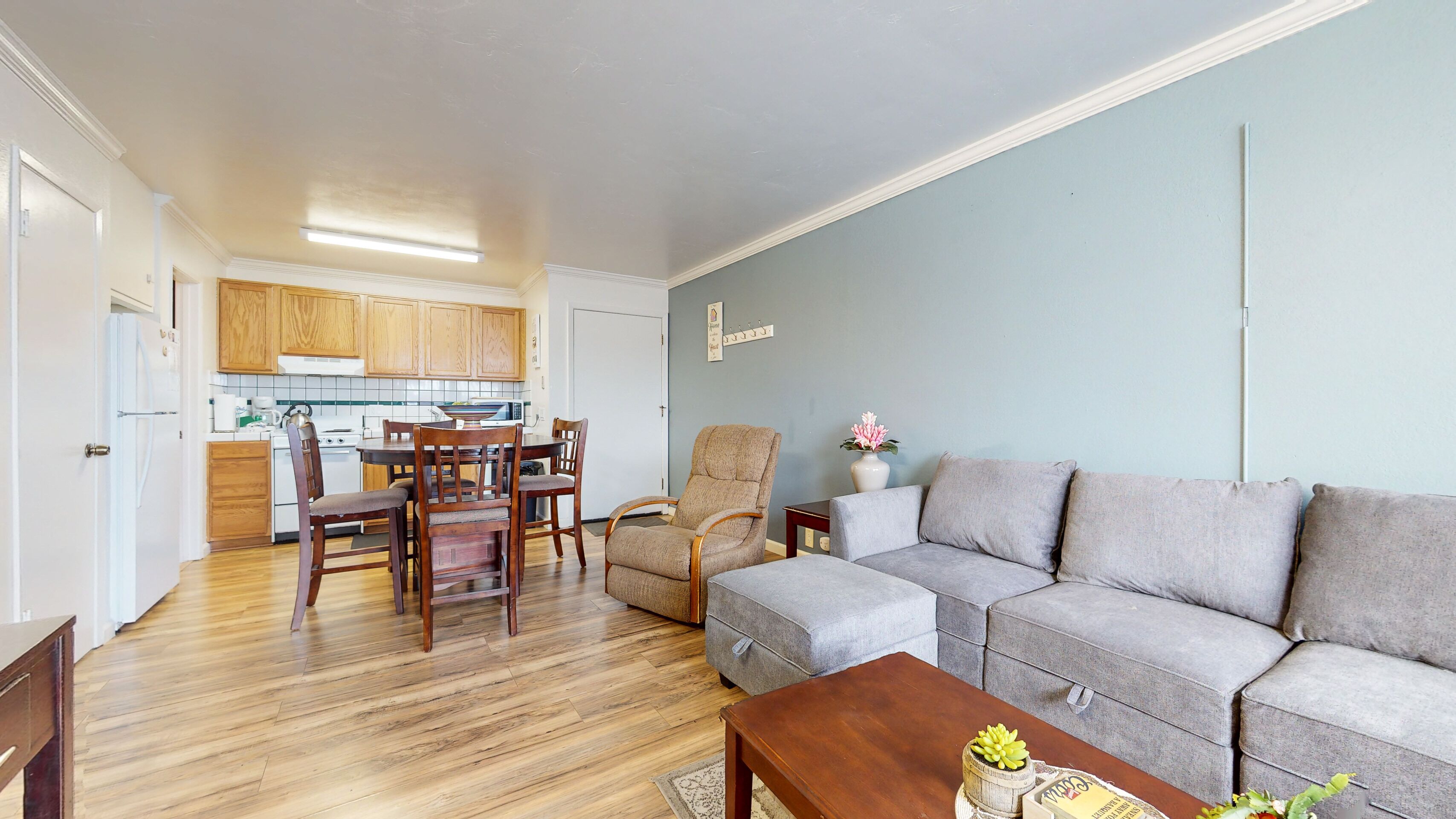 Een woonkamer met een grijze bank, een houten eettafel met stoelen, een grijze fauteuil en een klein bijzettafeltje met een vaas.