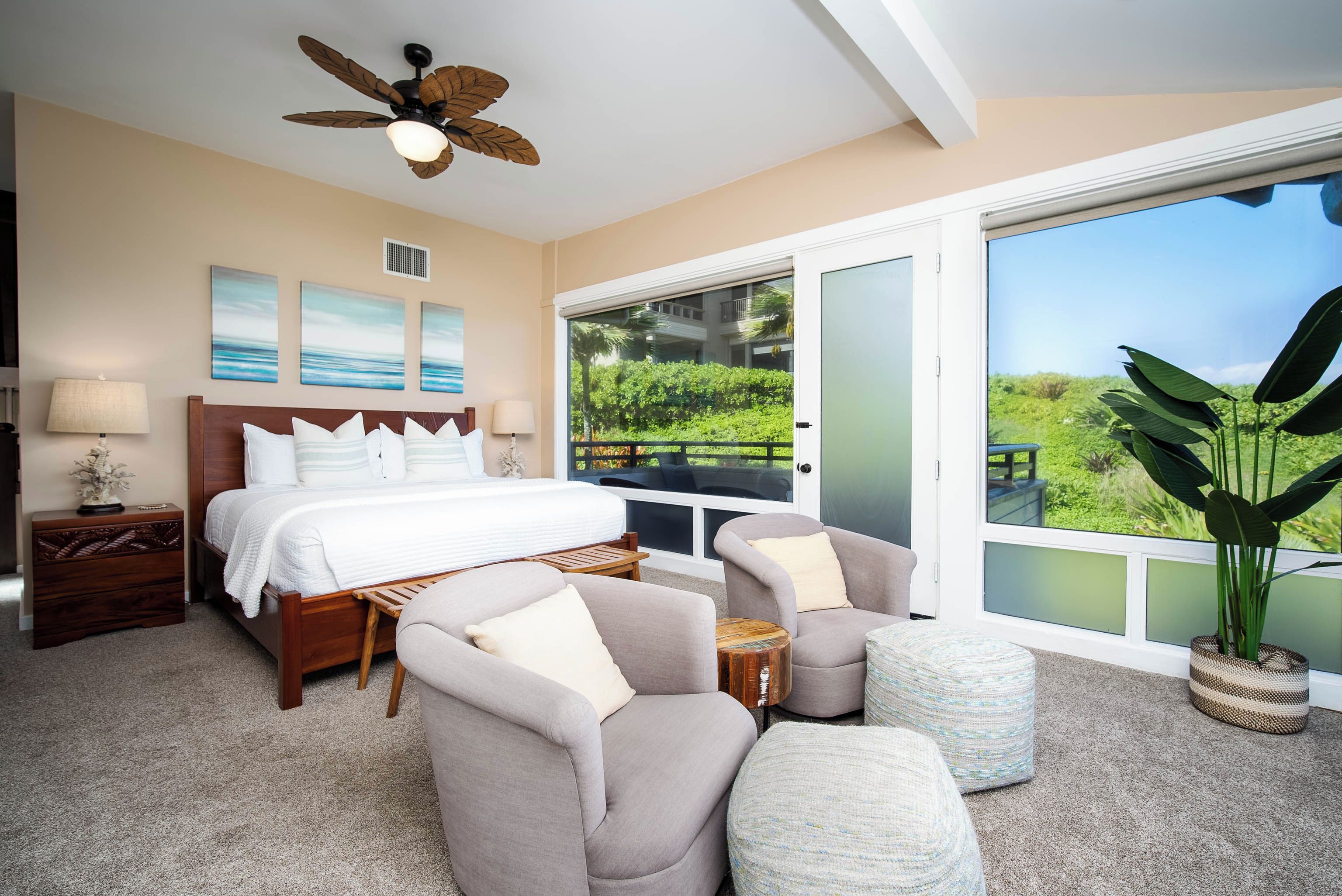 A bedroom with a bed, two armchairs, a side table, a ceiling fan, and a large window with a view of greenery.