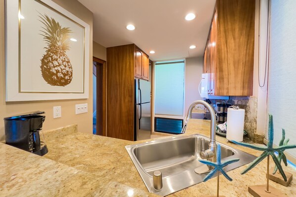 A kitchen with a stainless steel sink, wooden cabinets, a coffee maker, and a framed pineapple artwork.