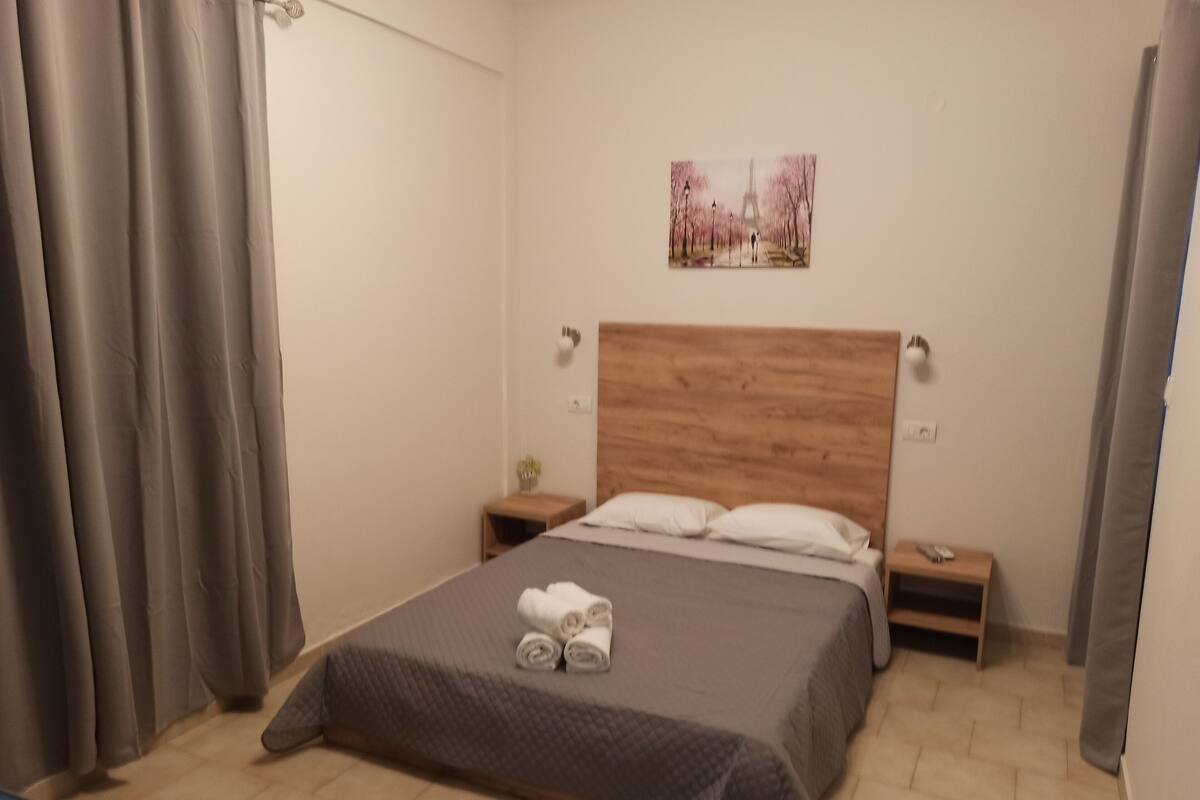 Um quarto com cabeceira de madeira, uma cama com colcha cinza, duas mesas de cabeceira com abajures e um quadro da Torre Eiffel.