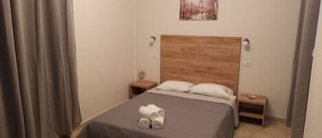 A bedroom with a wooden headboard, a bed with a grey quilt, two bedside tables with lamps, and a painting of the Eiffel Tower.