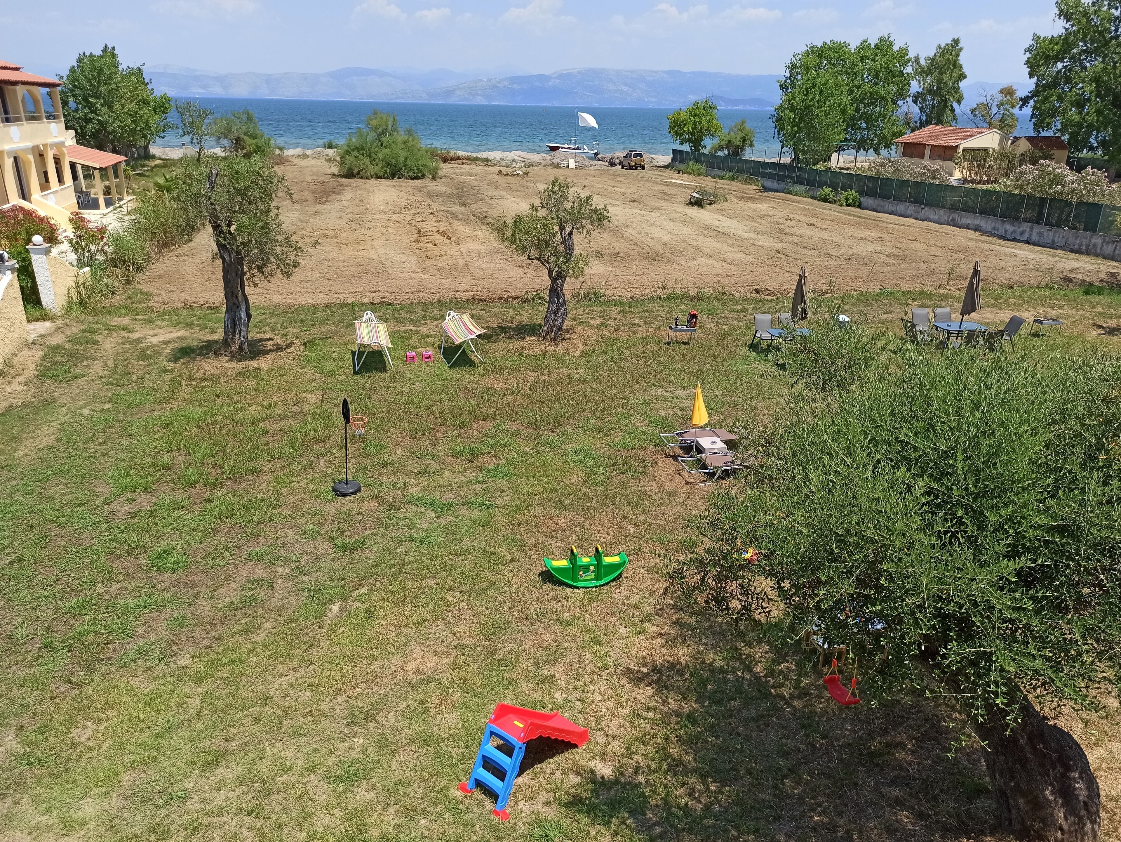 A grassy area with outdoor seating, a small boat, and a view of the sea and mountains in the background.
