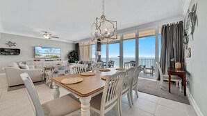 A spacious living room with a large TV, a dining area with a wooden table and white chairs, and a balcony with a view of the ocean.