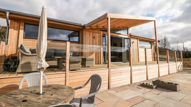 A modern wooden house with a patio area featuring wicker furniture and a table with an umbrella. - Stables Lodge (Chesterfield)
