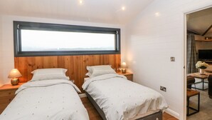 A bedroom with two beds, wooden headboards, a large window, and a small table with a chair. - Stables Lodge (Chesterfield)