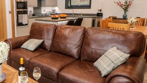 A brown leather sofa with two pillows, a bottle of wine, and two glasses on a wooden table. - Stables Lodge (Chesterfield)