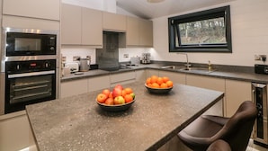 A modern kitchen with stainless steel appliances, a large island with fruit bowls, and a window with a view of the outdoors. - Stables Lodge (Chesterfield)