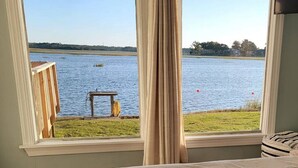 Une pièce dotée d’une grande fenêtre donnant sur un lac, d’une petite table avec une chaise et d’un lit recouvert d’une couverture à rayures.
