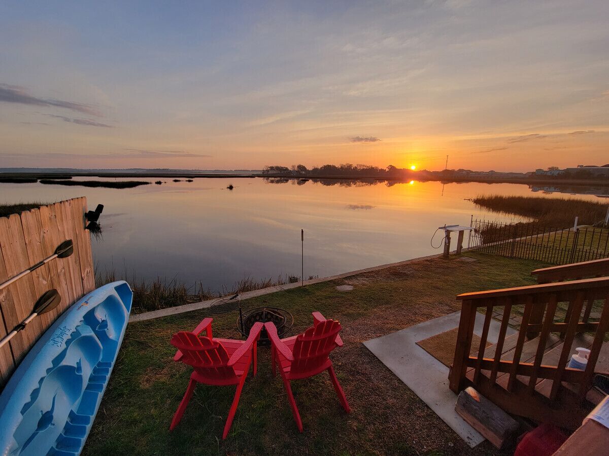 A lakeside view with a wooden deck, red Adirondack chairs, and a kayak.