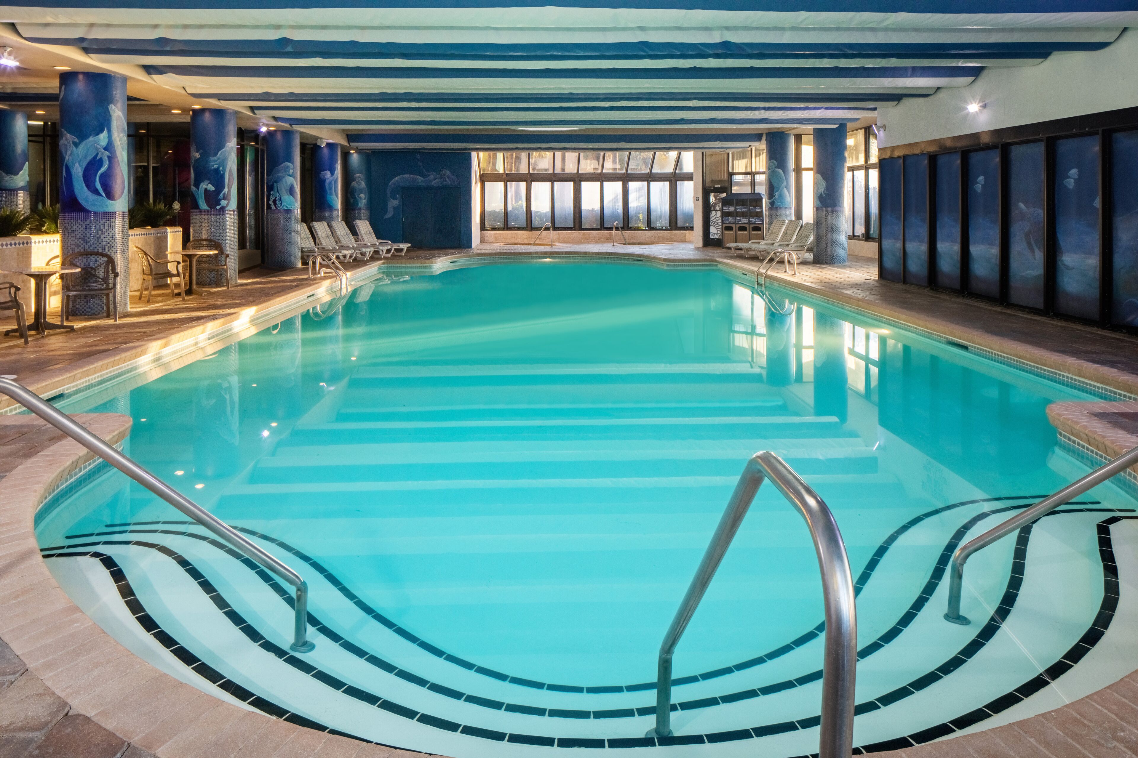 An indoor swimming pool with clear blue water, tiled steps leading into the pool, and lounge chairs along the sides.