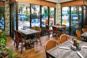 En restaurang med bord och stolar i trä, stora fönster med utsikt över en pool och palmer.