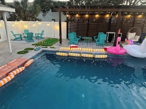 Une piscine avec des bouées en forme de flamants roses, des chaises vertes et une pergola en bois décorée de guirlandes lumineuses.